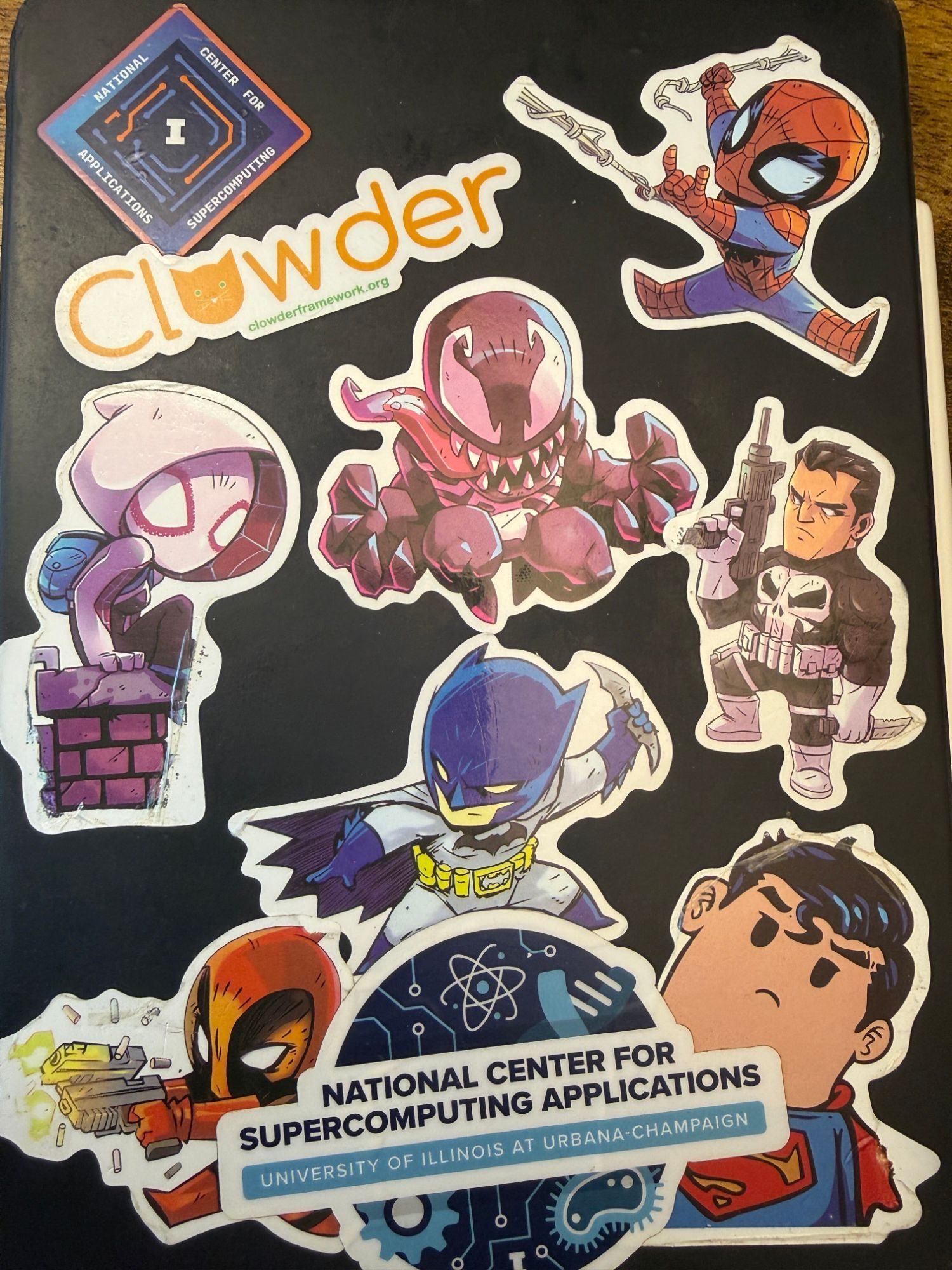 A dark laptop lid covered in stickers: NCSA and Clowder logos alongside chibi Spider-Man, Venom, Batman, Superman and other comic heroes